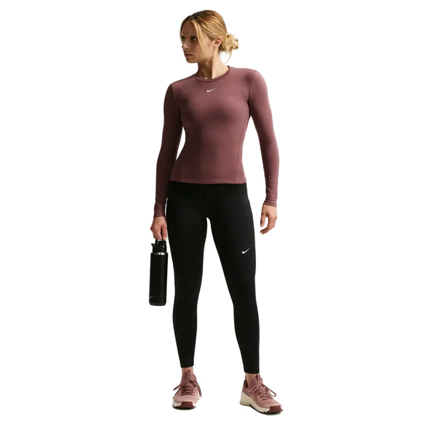 En kvinde i sportstøj holder en vandflaske iført en rødbrun langærmet top, pink Nike-sneakers og sort/hvide Nike Nk Df One Hr 7/8 Pkt Tght-leggings til kvinder. Hun kigger til venstre mod en hvid baggrund.