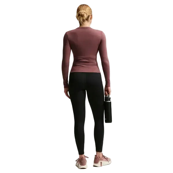 En person med blondt hår i en knold, iført en langærmet lilla top og Nike Nk Df One Hr 7/8 Pkt Tght Dame træningstights i sort/hvid samt pink sportssko, står med ansigtet vendt væk og holder en sort vandflaske i sin venstre hånd.
