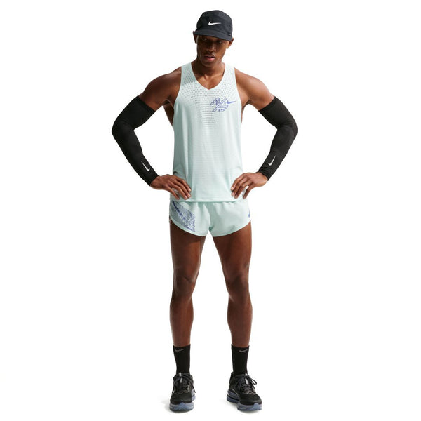 En mand står med ansigtet fremad og er iført Nike Nk Dfadv Aeroswift Gfx Snglt (herre) i Barely Green/Lapis, matchende shorts, sorte ærmer, sokker, løbesko og en kasket. Hans hænder hviler på hofterne mod en hvid baggrund.