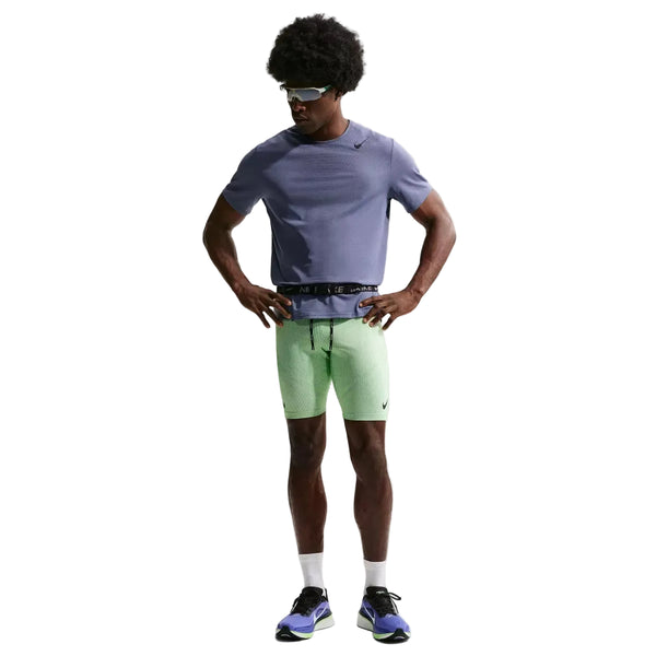 En person står med hænderne på hofterne iført en blå kortærmet skjorte, Nike AeroSwift Running Half Tights til mænd i Lime Blast/Steam/Light Silver/Black, solbriller, hvide sokker og lilla og sorte sportssko på en almindelig hvid baggrund.