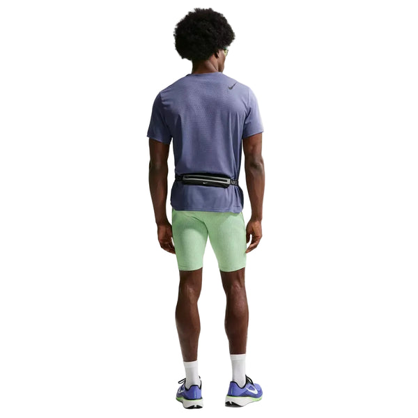 En person står med ryggen til og er iført en lilla trøje, Nike AeroSwift Running Half Tights til mænd i Lime Blast/Steam/Light Silver/Black, hvide sokker, blå og hvide sportssko og en lille sort bæltetaske på en almindelig hvid baggrund.