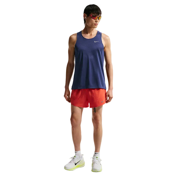 En person i Nike Nk Dfadv Stride Tank til mænd (Midnight Navy/Reflective Silver), røde shorts, hvide sokker, hvide løbesko med neongule såler og rødtonede solbriller står og kigger lidt til siden.