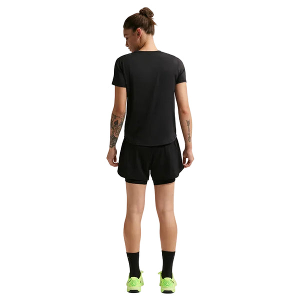 En person med knold står med ansigtet væk på en almindelig hvid baggrund, viser tatoveringer på armen og er iført en sort t-shirt, Nike Nk One Df Hr 3In 2N1 Short Dame i sort/hvid, sorte sokker og neongrønne sneakers.