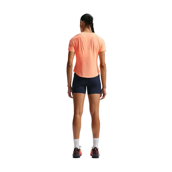 En kvinde med flettet hår kigger væk fra kameraet iført en Nike Nk Swft Nvlt Brthe Ss Top (Dame, Orange Pulse/Photon Dust/Reflective Silv), navyfarvede shorts, hvide sokker og sorte sportssko - ideelt til løbetræning af kvinder.