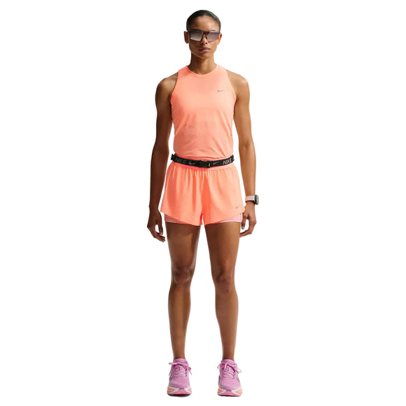 En kvinde kigger fremad i en Nike Nk Swift Nvlt Brthe Tank, Dame, Orange Pulse/Photon Dust/Reflective Silv med matchende shorts, pink løbesko, sort taljebælte, solbriller og et smartwatch. Den livlige tank popper op mod den hvide baggrund.