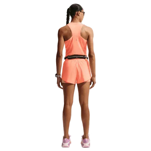 En person med flettet hår kigger væk og er iført en Nike Nk Swift Nvlt Brthe Tank til kvinder i Orange Pulse/Photon Dust/Reflective Silv, matchende shorts, et sort bælte, solbriller, et armbåndsur og lyserøde løbesko.