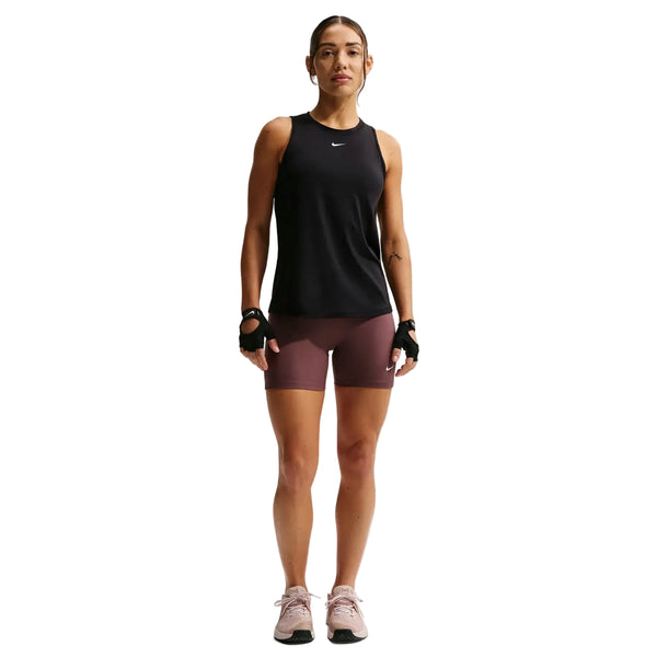 En kvinde står med front mod Nike træningstøj dame: en sort/hvid Nike Nk One Classic Df Tank, rødbrune shorts, fingerløse handsker og pink sneakers på en hvid baggrund.