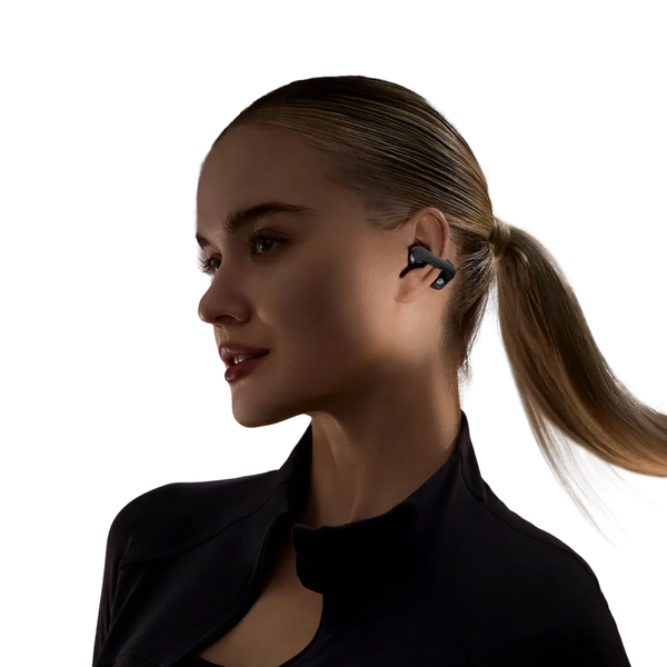 En kvinde med blondt hår i hestehale bærer sorte Shokz Open Dots One True Wireless Open-Ear Hovedtelefoner og en sort top med lynlås, mens hun kigger lidt til siden mod en almindelig hvid baggrund.