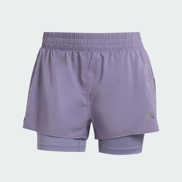 Adidas Otr B S 2In1 dame løbeshorts i lys lilla med elastisk foring og dobbeltlag. Svedtransporterende materiale gør dem perfekte til træning. Adidas logoet ses ved kanten, og baggrunden er ensfarvet og lys.
