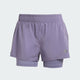 Adidas Otr B S 2In1 dame løbeshorts i lys lilla med elastisk foring og dobbeltlag. Svedtransporterende materiale gør dem perfekte til træning. Adidas logoet ses ved kanten, og baggrunden er ensfarvet og lys.