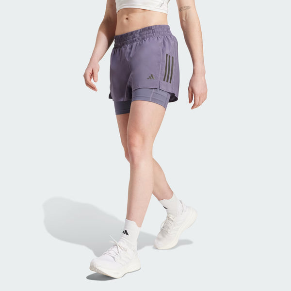 En person iført en hvid crop top og lilla Adidas Otr B S 2In1, Dame, Previo shorts med sorte striber, parret med hvide sokker og sneakers, går fremad mod en almindelig lys baggrund.