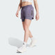 En person iført en hvid crop top og lilla Adidas Otr B S 2In1, Dame, Previo shorts med sorte striber, parret med hvide sokker og sneakers, går fremad mod en almindelig lys baggrund.