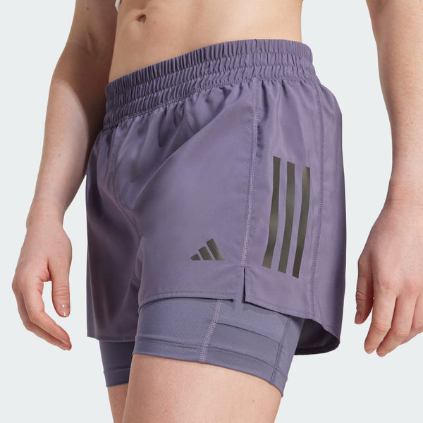 En person er iført Adidas Otr B S 2In1 Dame Previo lilla løbeshorts med sorte striber over matchende kompressionsshorts. Kun underkroppen og overbenene er synlige mod en ensfarvet baggrund.