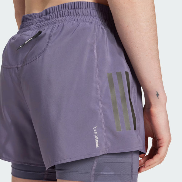 En person er iført Adidas Otr B S 2In1 dame løbeshorts i lyslilla med en lille lynlåslomme, tre lodrette refleksstriber og Climacool-tekst på siden. På venstre håndled er der tatoveret et lille lyn.
