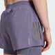 En person er iført Adidas Otr B S 2In1 dame løbeshorts i lyslilla med en lille lynlåslomme, tre lodrette refleksstriber og Climacool-tekst på siden. På venstre håndled er der tatoveret et lille lyn.
