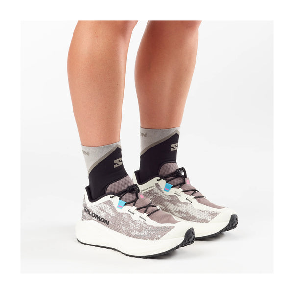 En person iført sorte og grå sokker og Salomon Aero Glide 4 Grvl Dame-sko i Vanilla Ice/Black/Iron står på en hvid baggrund. Kun underbenene og fødderne er synlige.