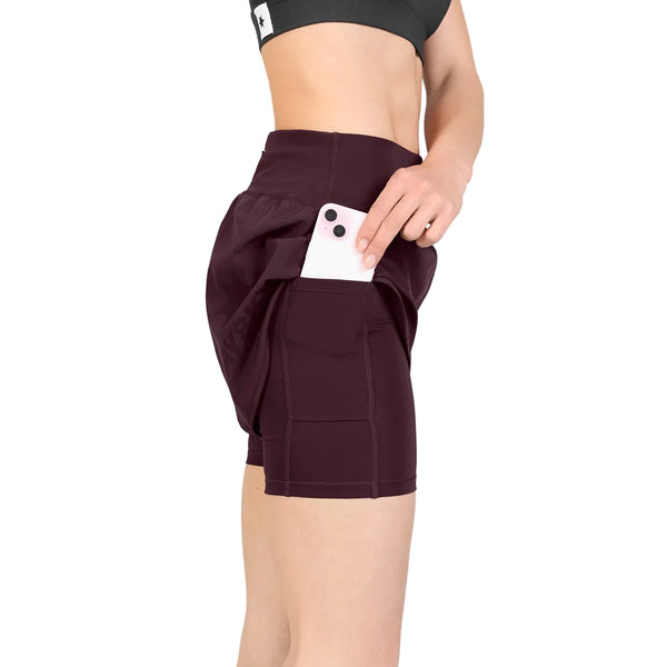 En kvinde iført en sort sports-BH og et par Saysky Pace 2-i-1-shorts (3″, Dame) i farven burgundy lægger en hvid smartphone i shortsens sidelomme. Baggrunden er ensfarvet hvid.