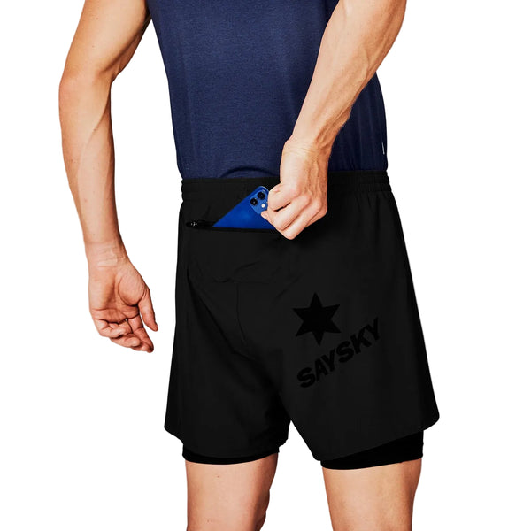 En person i Saysky Pace 2in1 Running Shorts 5", Herre, Black og en marineblå top lægger en blå smartphone i shortsens baglomme med lynlås. "SAYSKY" og et stjernelogo er trykt på de sorte herre-shorts.