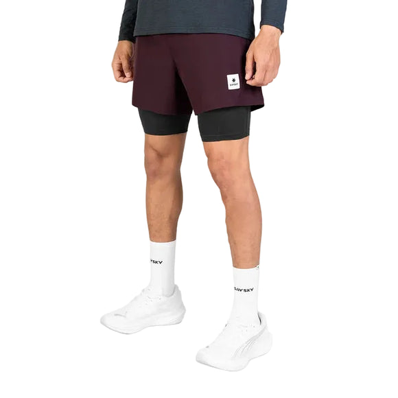 Der vises et billede af en person fra livet og ned, iført Saysky Pace 2-i-1-shorts i størrelse 5″ i farven burgundy over sorte kompressionsshorts, hvide strømper med teksten »TW SKY« og hvide sportssko.