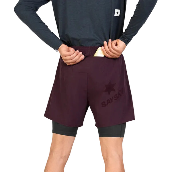 Iført en langærmet trøje og et par Saysky Pace 2-i-1-shorts 5″ til mænd i farven burgundy samt sorte underbukser lægger en person (hvis hoved ikke er synligt) en telefon eller et kort i baglommen på løbeshortsene.