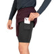 En person stikker en smartphone ned i sidelommen på et par sorte kompressionsshorts, der bæres under et par Saysky Pace 2in1 Shorts 5″, Herre, Burgundy. Kun den nederste del af overkroppen og lårene er synlige, og personen har desuden en mørk langærmet trøje på.