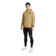 En person står med ansigtet vendt fremad iført en beige Saysky Pace Tech-jakke til mænd med hætte, kombineret med sorte leggings med et lille rektangulært mærke og hvide sportssko, foran en ensfarvet hvid baggrund.