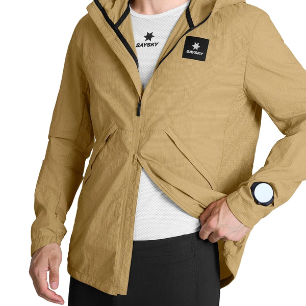 En person, der bærer en beige Pace Tech-jakke fra Saysky over en hvid mesh-trøje, retter på den funktionelle træningsjakke med venstre hånd, mens vedkommende også har et sort armbåndsur på venstre håndled.