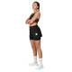 En person med flettet hår står med armene over kors, iført en sort sports-BH, Saysky Pace 2-i-1-shorts 3″ Dame i sort, hvide strømper og hvide sneakers foran en ensfarvet lysegrå baggrund.