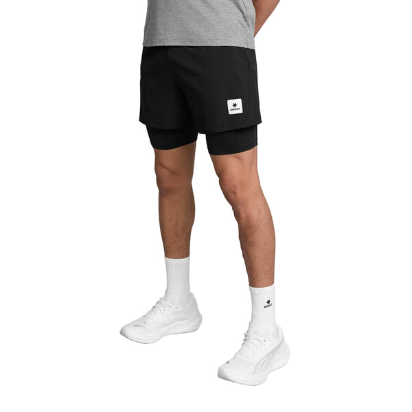 En person, der står op og er iført en grå T-shirt, Saysky Pace 2-i-1-shorts 5″, Herre, i sort, hvide strømper og hvide sportssko. Billedet er beskåret ved den øverste del af brystet, så personens hoved og overkrop ikke er synlige.