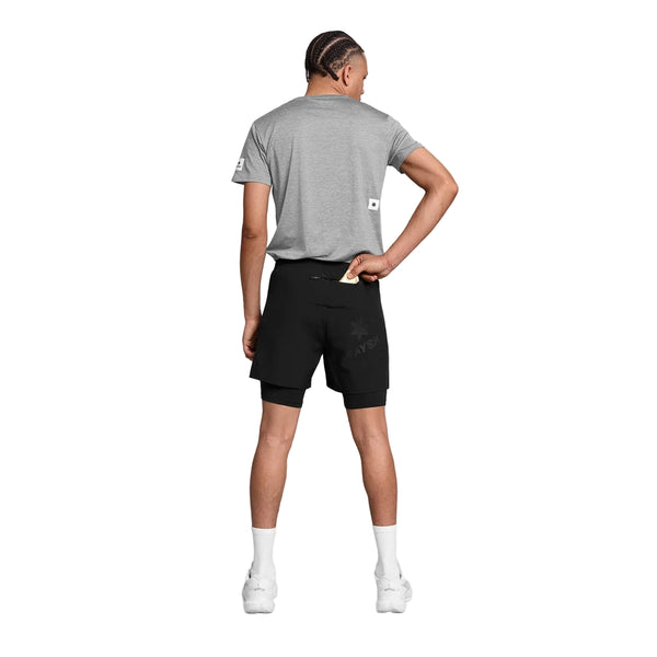 En person står med ryggen til kameraet foran en ensfarvet hvid baggrund. Personen er iført en grå T-shirt, et par Saysky Pace 2-i-1-shorts 5″ til mænd i sort – designet som trail-løbeshorts – hvide sokker og hvide sko, med den ene hånd på hoften og den anden hånd nede i lommen på shortsene.