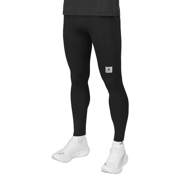 En person har Saysky Pace+ lange herretights i sort på, med et diskret logo på låret, og er klædt i hvide sneakers og en mørk skjorte. Billedet er beskåret over taljen, så det kun viser den nedre del af overkroppen, benene og fødderne.