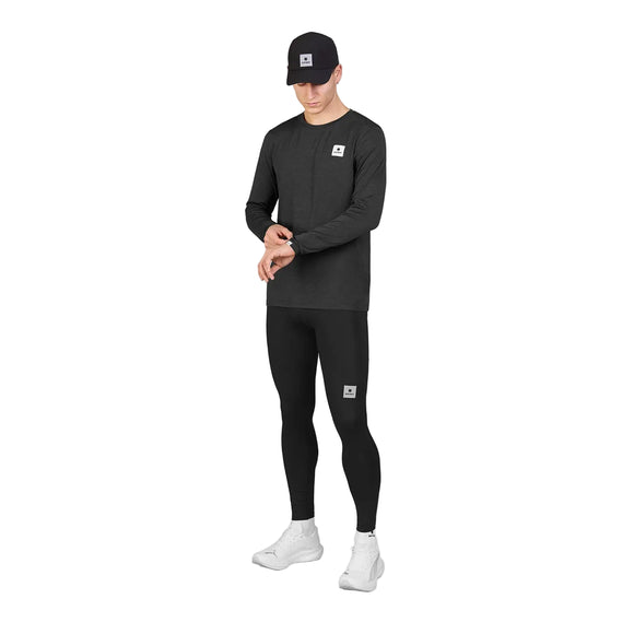 En person iført en sort kasket, en sort langærmet trøje, Saysky Pace+ Long Tights til herre i sort og hvide sneakers står foran en hvid baggrund og kigger ned, mens vedkommende retter på ærmet. Hvert stykke tøj er forsynet med et lille firkantet Saysky-logo.