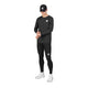 En person iført en sort kasket, en sort langærmet trøje, Saysky Pace+ Long Tights til herre i sort og hvide sneakers står foran en hvid baggrund og kigger ned, mens vedkommende retter på ærmet. Hvert stykke tøj er forsynet med et lille firkantet Saysky-logo.