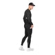 Der ses en person, der står i profil foran en ensfarvet hvid baggrund, iført Saysky Pace+ Long Tights, Herre, i sort. Personen har desuden en sort langærmet trøje, en sort hat og hvide sneakers på og holder en lyserød telefon i højre hånd.
