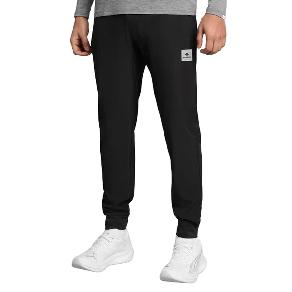 En person iført en grå langærmet skjorte og sorte unisex Pace-bukser fra Saysky med diskrete refleksdetaljer, kombineret med hvide sneakers, står foran en ensfarvet hvid baggrund.