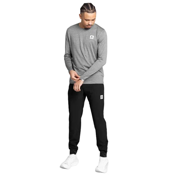 En person står foran en ensfarvet hvid baggrund iført en grå unisex-trøje med lange ærmer, Saysky Pace-bukser (unisex, sorte) og hvide sneakers. Personen kigger ned og retter sin venstre ærme med højre hånd.