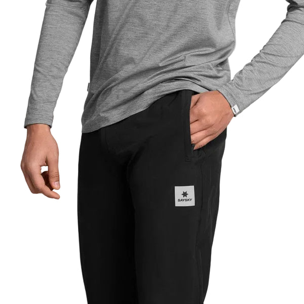En person iført en grå langærmet skjorte og de sorte unisex-bukser »Pace Pants« fra Saysky, med et SAYSAY-logo-mærke på lommen, står med den ene hånd i lommen foran en ensfarvet baggrund.