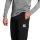 En person iført en grå langærmet skjorte og de sorte unisex-bukser »Pace Pants« fra Saysky, med et SAYSAY-logo-mærke på lommen, står med den ene hånd i lommen foran en ensfarvet baggrund.