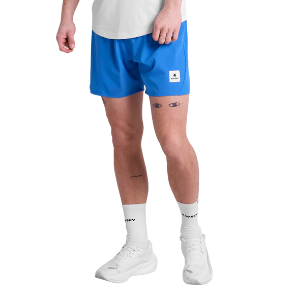 En person er iført en hvid t-shirt, Saysky Pace Shorts 5" Herre i blå med en lille logopatch, hvide sokker med sort tekst og hvide sneakers; på benene ses flere små tatoveringer som øjne og en bølge.