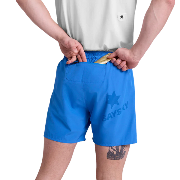 En person i hvid skjorte og blå Saysky Pace Shorts 5" Herre ses bagfra i færd med at stikke en guldindpakket genstand ned i baglommen på performance-shortsene, som bærer ordet "herre" og et stjernelogo.