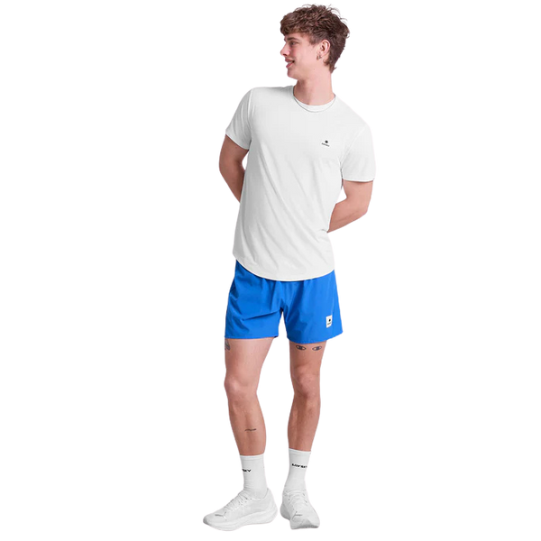 En person står på en almindelig baggrund iført en hvid t-shirt, Saysky Pace Shorts 5" Herre i blå, hvide sokker og hvide sneakers, med hænderne foldet bag ryggen og kigger til siden.