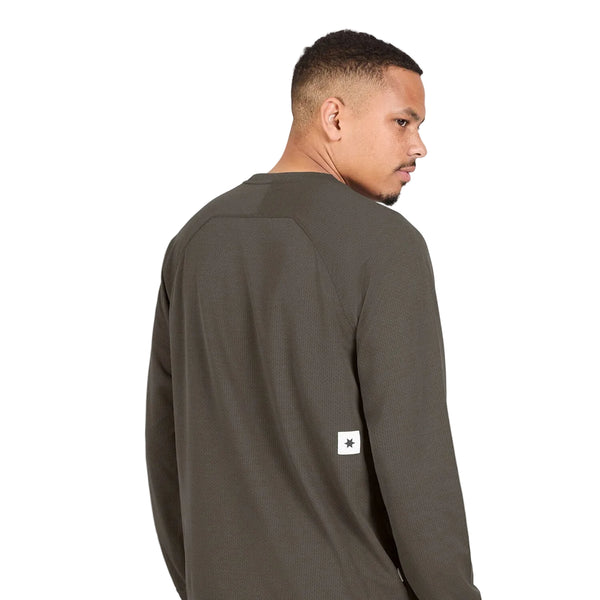 En mand med kort hår står med ryggen til kameraet og har den brune Pace Tech Long Sleeve, Herre fra Saysky på. Den langærmede løbetrøje er i en mørk olivenbrun nuance og fremstillet af åndbart materiale, med et lille hvidt, rektangulært mærke med en sort stjerne nederst på ryggen. Trøjen er udstyret med SAYSKYDRY-teknologi for øget komfort. Baggrunden er ensfarvet hvid.