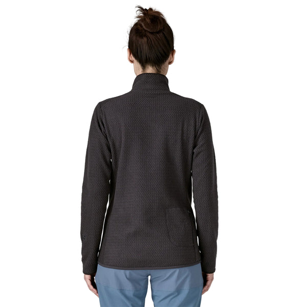 En kvinde med brunt hår i en knold ses bagfra iført Patagonia R1 Air Zip Neck fleecejakke i sort og lyseblå bukser på en hvid baggrund.