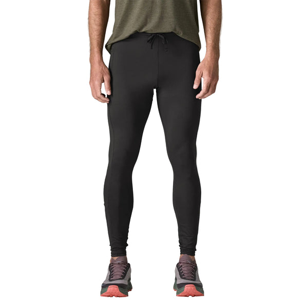 En person iført en grøn kortærmet T-shirt, Peak Mission Tights, Herre i sort fra Patagonia og grå løbesko med røde såler står foran en ensfarvet hvid baggrund. Kun kroppen fra skuldrene og ned er synlig.