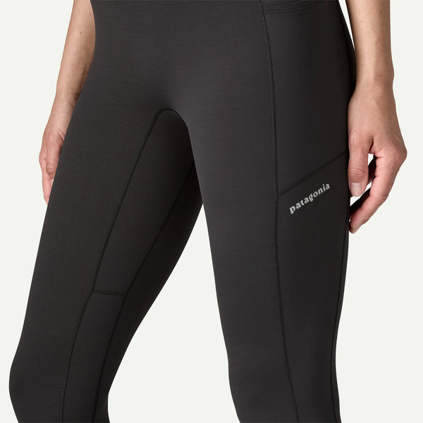 En person iført sorte Patagonia Peak Mission Tights 27" til kvinder vises fra taljen til overbenene. Strømpebukserne har en synlig sidelomme på låret med Patagonia-logoet over.
