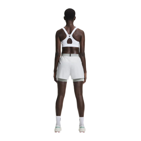 En person kigger væk iført On Performance Bra, Dame, White - perfekt til løb eller sportslige aktiviteter - sammen med lyse sportsshorts, hvide sokker og sportssko på en almindelig hvid baggrund.