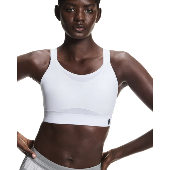 En person iført On Performance Bra, Dame, White med grå sportsbukser står selvsikkert på en almindelig hvid baggrund, klar til løb eller træning.