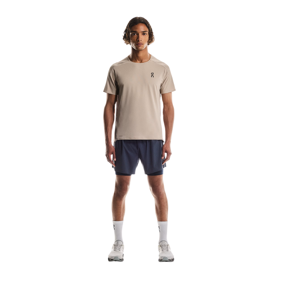 En mand ser frem for sig iført en On Performance-T til mænd i Desert/Cinder, marineblå shorts, hvide strømper og hvide løbesko, mens han står på en almindelig hvid baggrund.