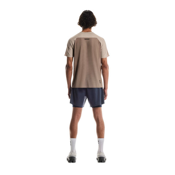 En person står med front mod kameraet iført en On Performance-T til mænd i farven Desert/Cinder, sammen med mørkeblå shorts, hvide sokker og lyse sportssko på en hvid baggrund.
