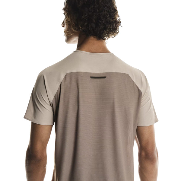 En person med krøllet hår ses bagfra iført On Performance-T, Herre, Desert/Cinder - en åndbar træningstrøje i lys beige med et mørkere beige panel og en lille rektangulær detalje på den øverste del af ryggen for optimal komfort.
